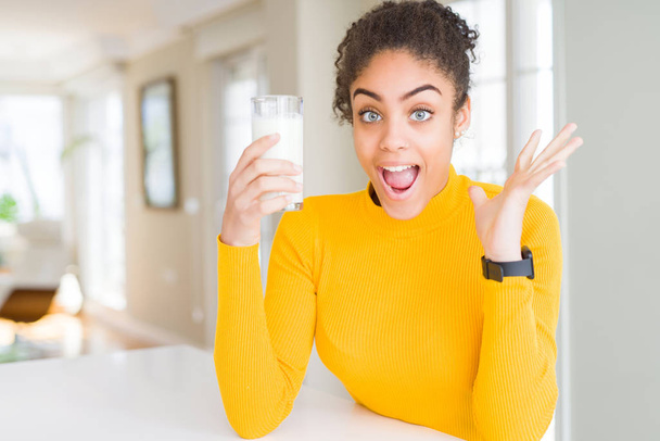 Jonge african american vrouw het drinken van een glas verse melk zeer blij en opgewonden, winnaar expressie vieren overwinning schreeuwen met grote glimlach en getogen handen - Foto, afbeelding