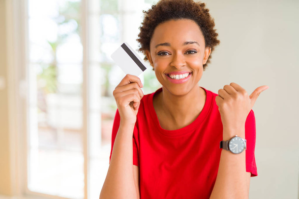 Młody african american kobieta trzyma karty kredytowej, wskazując i Wyświetlono z kciukiem do boku z szczęśliwy twarz uśmiechający się - Zdjęcie, obraz