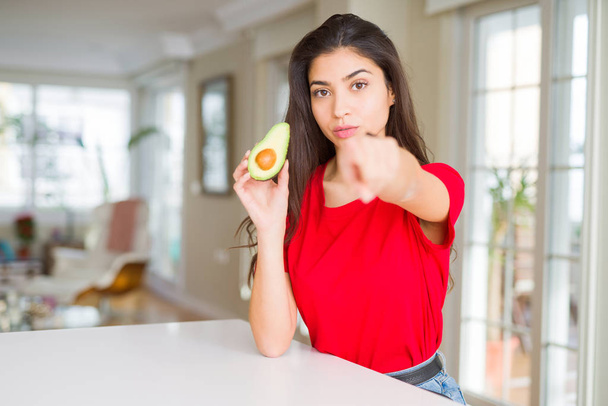 Genç kadın sağlıklı avokado ile kamera ve size, el işareti, pozitif ve kendinden emin bir jest ön parmakla yeme - Fotoğraf, Görsel
