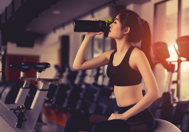 Asian woman drinking protein shake from bottle after workout exercise for muscle at fitness gym healthy lifestyle bodybuilding sitting on fitness ball - Foto, Bild