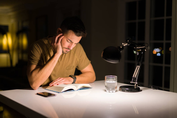 Young handsome man studying at home, reading a book at night - Photo, Image