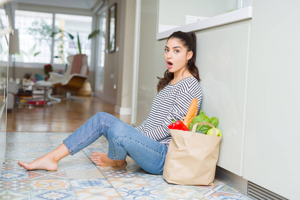 Młoda kobieta siedzi na podłodze w kuchni z papieru torbą pełną świeże Artykuły spożywcze, boi się i wstrząśnięty wyrażenie zdziwienie, strach i podekscytowana twarz. - Zdjęcie, obraz