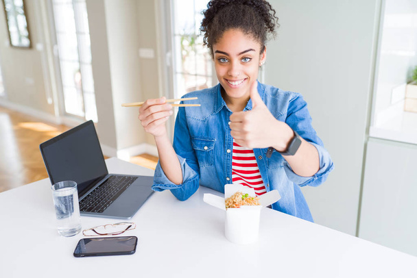 Joven mujer de negocios afroamericana trabajando y comiendo fideos asiáticos feliz con una gran sonrisa haciendo signo de ok, pulgar hacia arriba con los dedos, señal excelente
 - Foto, imagen