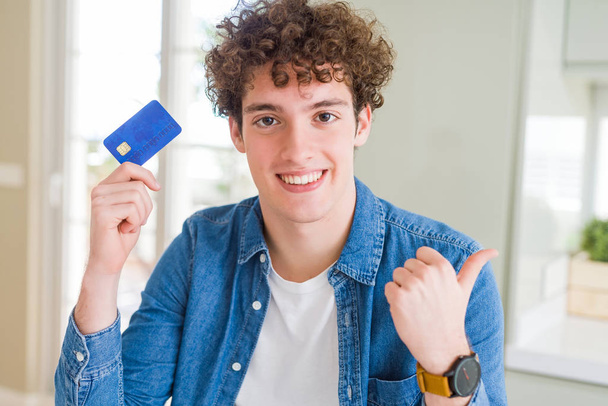 Jonge man met creditcard aan te wijzen en te tonen met de duim aan de kant met een blij gezicht lachend - Foto, afbeelding
