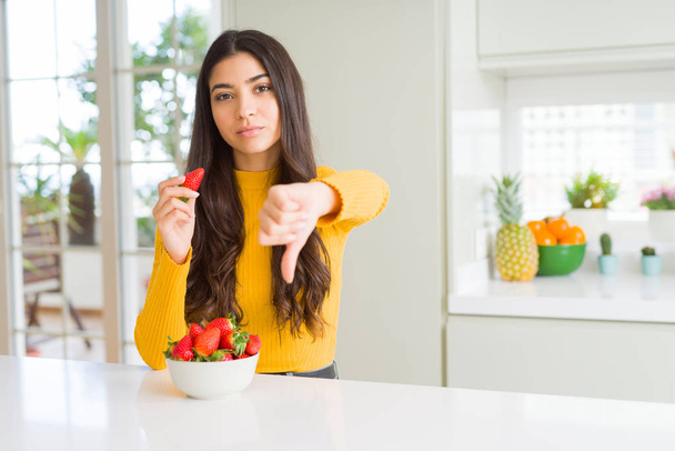 Mujer joven comiendo fresa roja fresca como bocadillo saludable con la cara enojada, signo negativo que muestra aversión con los pulgares hacia abajo, concepto de rechazo
 - Foto, imagen