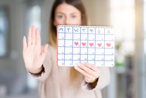 Молода красива жінка тримає календар менструацій вдома з відкритою рукою робить знак зупинки з серйозним і впевненим виразом, жест захисту
 - Фото, зображення