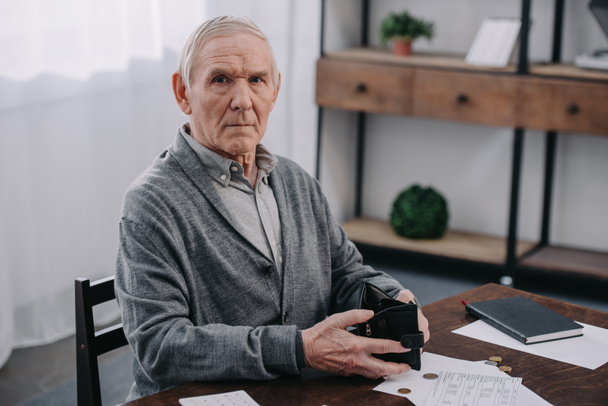 male pensioner sitting at table with paperwork, looking at camera and holding wallet  - Foto, Bild