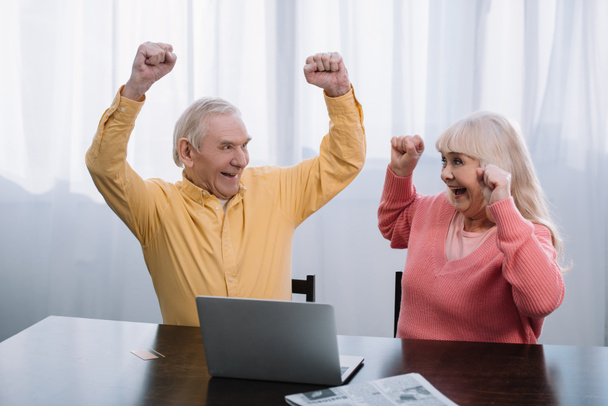 szczęśliwa para starszy w kolorowe ubrania, siedział przy stole z laptopa i doping z ręce w powietrzu - Zdjęcie, obraz