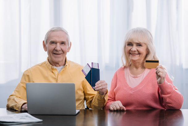 Senior paar met vliegtickets, paspoorten en creditcard siiting aan tafel met laptop en de camera kijken - Foto, afbeelding