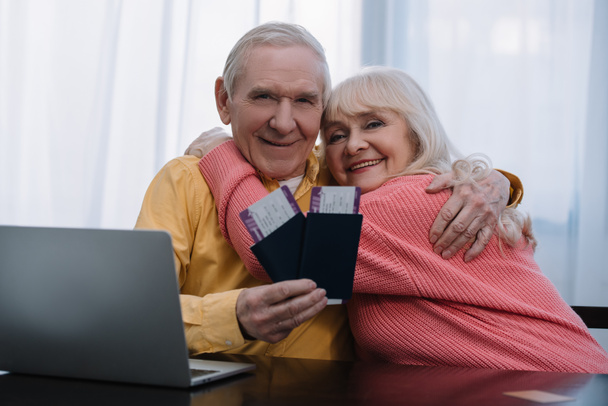 heureux couple de personnes âgées assis à table avec ordinateur portable, étreignant et tenant des billets d'avion avec passeports à la maison
  - Photo, image