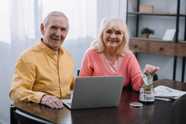 senior couple sitting at table with laptop and looking at camera while woman putting money in glass jar with 'pension' lettering - Photo, Image