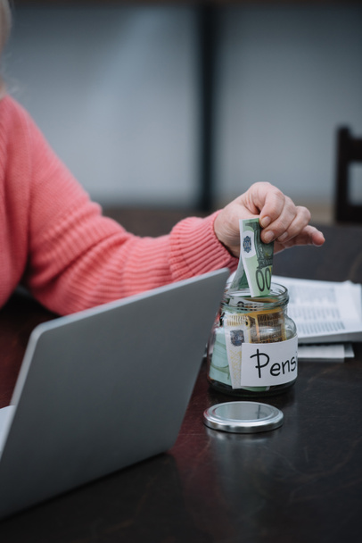 обрезанный вид пожилой женщины, сидящей за столом с ноутбуком, в то время как положить деньги в стеклянную банку с "пенсионного" слова
 - Фото, изображение