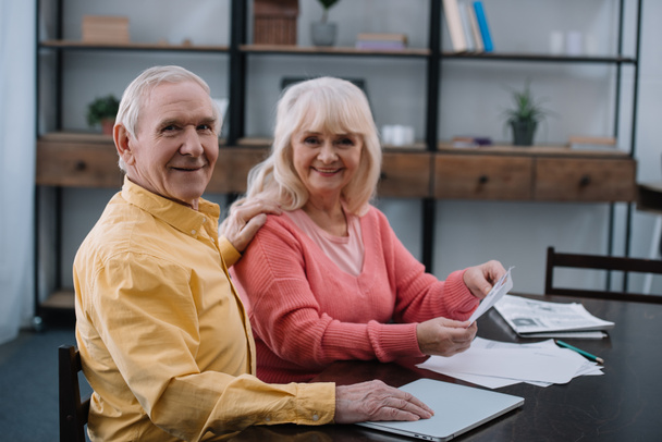 glückliches Seniorenpaar blickt in die Kamera, während es mit Dokumenten am Tisch sitzt - Foto, Bild