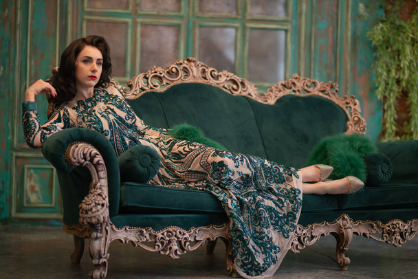 menina caucasiana elegante em um vestido de renda de lantejoulas longas de luxo com uma boa fofa verde em suas mãos posando no quarto retro com mobiliário de luxo vintage
 - Foto, Imagem