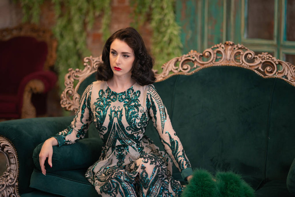 elegant caucasian girl in a luxurious long sequins lace dress with a green fluffy boa in her hands posing in the retro room with vintage luxury furniture - Photo, Image