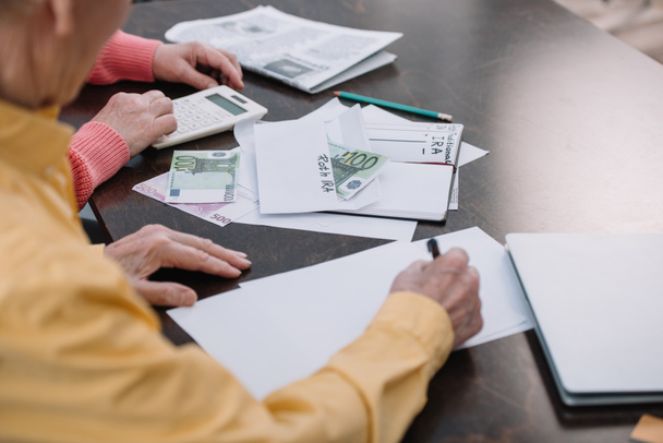 Seniorenpaar sitzt mit Papierkram am Tisch, Umschlag mit "roth ira" -Schriftzug und Geld - Foto, Bild
