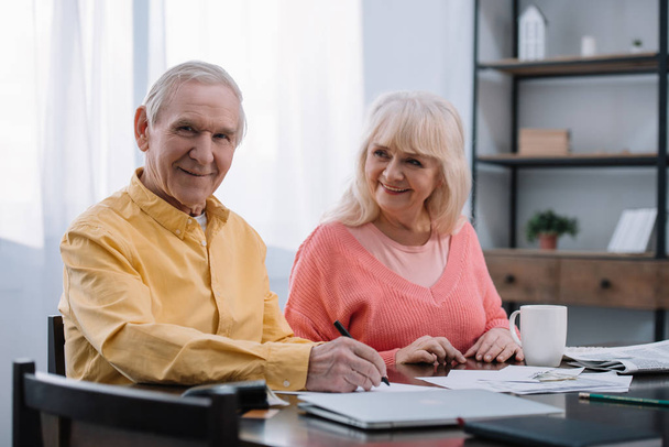щаслива старша пара сидить за столом і заповнює документи вдома
 - Фото, зображення