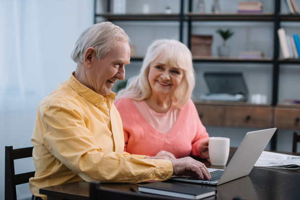 senior couple in colorful clothes sitting at table with coffee cup and using laptop - Fotó, kép