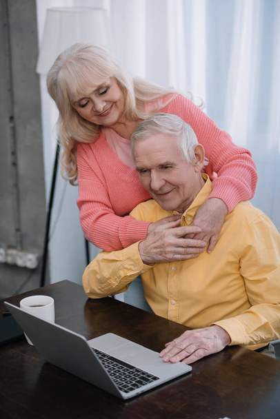 sonriendo pareja de ancianos abrazándose mientras el hombre sentado en la mesa y el uso de la computadora portátil
 - Foto, Imagen