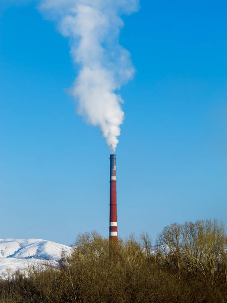 Spessa fumo bianco esce dalla pipa rossa della fabbrica contro un cielo blu chiaro, montagne innevate e cime degli alberi
. - Foto, immagini