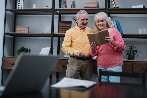 selective focus of happy senior couple in colorful clothes reading book in living room - Fotó, kép