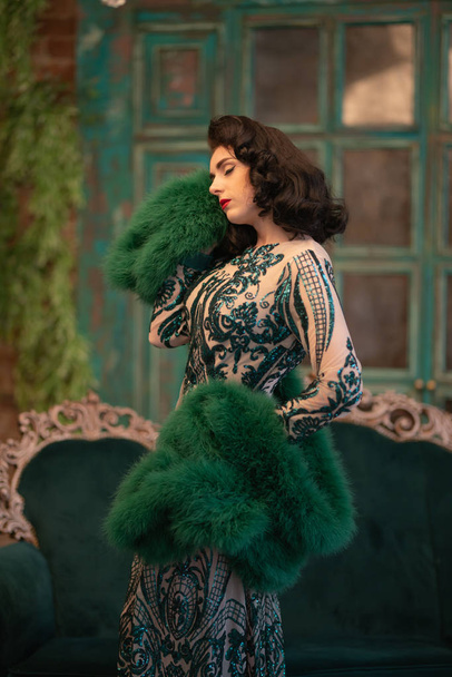 elegant caucasian girl in a luxurious long sequins lace dress with a green fluffy boa in her hands posing in the retro room with vintage luxury furniture - Photo, image