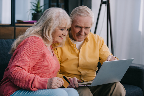 lachende senior paar zittend op de Bank met laptop en creditcard terwijl het doen van online winkelen bij u thuis - Foto, afbeelding