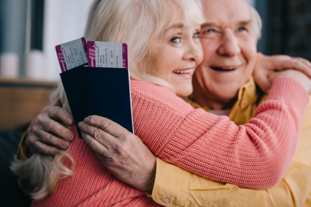 smiling senior couple hugging and holding air tickets with passports at home  - Photo, Image