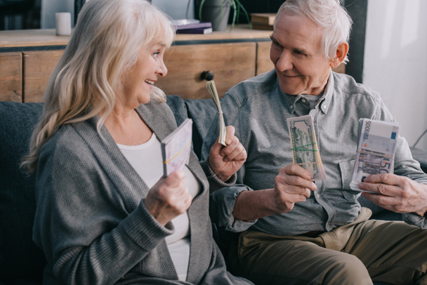 glückliches Seniorenpaar auf Couch sitzend und Euro- und Dollarscheine in der Hand - Foto, Bild