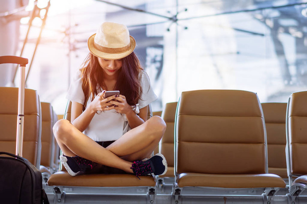 Asian woman teenager using smartphone at airport terminal sitting with luggage suitcase and backpack for travel in vacation summer relaxing waiting flight transport - Photo, image