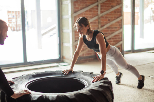 Young trained woman working out with truck tire with personal trainer assistance - Photo, Image