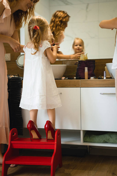 Mom and daughters do makeup in the bathroom, apply lipstick in front of the mirror. Mom and daughters have fun, spend time together. - Fotografie, Obrázek