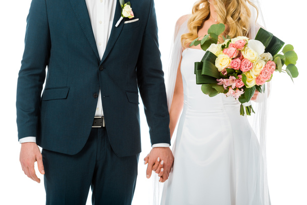 περικοπεί άποψη της νύφης με όμορφη γαμήλια ανθοδέσμη, κρατώντας τα χέρια με το γαμπρό στο κομψό κοστούμι που απομονώνονται σε λευκό - Φωτογραφία, εικόνα