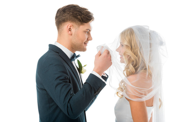 noivo feliz olhando para o rosto de noivas sorridentes enquanto levanta véu nupcial isolado no branco
 - Foto, Imagem