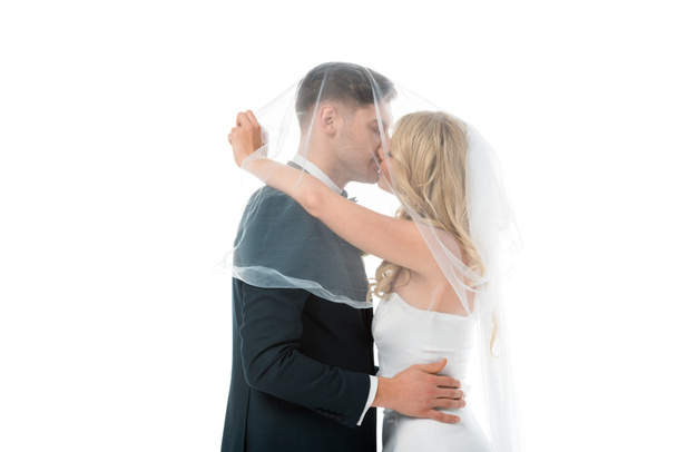 happy bride kissing bridegroom while covering him with bridal veil isolated on white - Foto, imagen