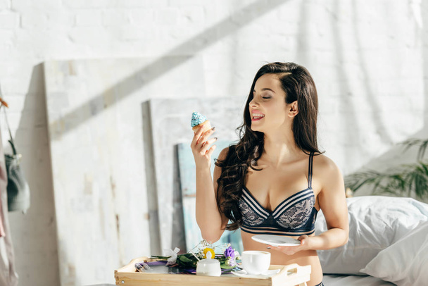 cheerful woman in underwear holding tasty cupcake near tray with breakfast  - Zdjęcie, obraz
