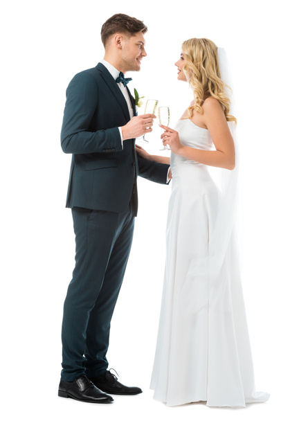 cheerful bride and groom clinking glasses of champagne and looking at each other isolated on white - Foto, Imagem