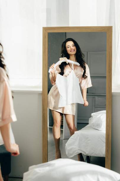 κρεμάστρα εκμετάλλευση ελκυστική μελαχρινή γυναίκα με ρούχα ενώ στέκονται κοντά καθρέφτη - Φωτογραφία, εικόνα