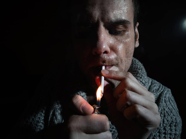 Dark and sullen picture of a young man Smoking on a black background. A man lights a cigarette - Фото, зображення