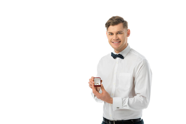 usmívající se ženich drží krabičky s snubní prsten a při pohledu na fotoaparát izolované na bílém - Fotografie, Obrázek