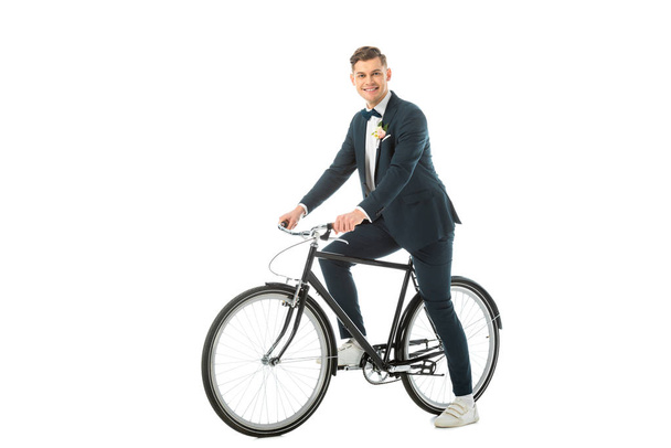 cheerful handsome bridegroom in elegant suit and sneakers sitting on bike isolated on white - Φωτογραφία, εικόνα
