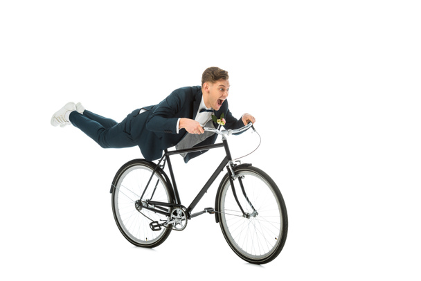 ενθουσιασμένος γαμπρός στο κομψό κοστούμι κάνει ακροβατικά με ποδήλατο που απομονώνονται σε λευκό - Φωτογραφία, εικόνα