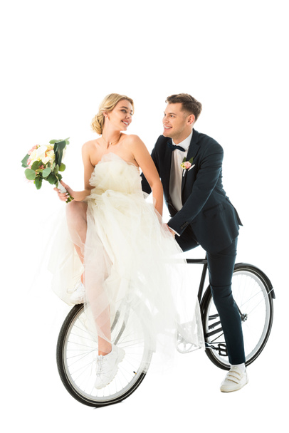 ευτυχισμένη νύφη με γαμήλια ανθοδέσμη κάθεται στο ποδήλατο μαζί με χαμόγελο γαμπρός απομονωθεί σε λευκό - Φωτογραφία, εικόνα