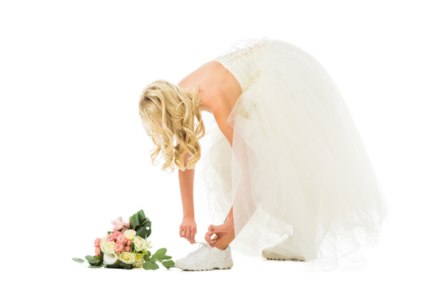 beautiful bride in wedding dress tying shoelaces on sneakers isolated on white - Photo, Image