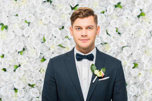 handsome groom in elegant jacket with boutonniere looking at camera on white floral background - Фото, изображение
