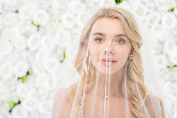 portrait of beautiful young bride with face covered with transparent bridal veil on white floral background - Foto, Bild