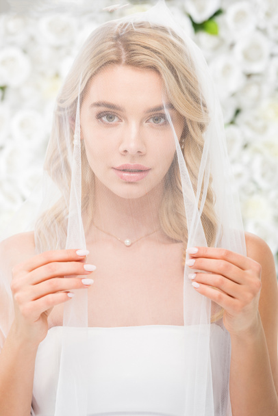 schöne junge Frau mit Gesicht, das mit transparentem Brautschleier bedeckt ist und in die Kamera auf weißem Blumenhintergrund blickt - Foto, Bild