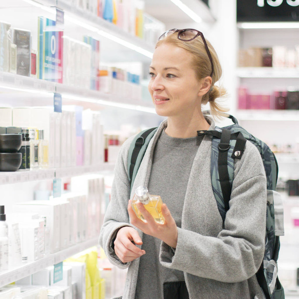 Blond young female traveler wearing travel backpack choosing perfume in airport duty free store. - Photo, Image