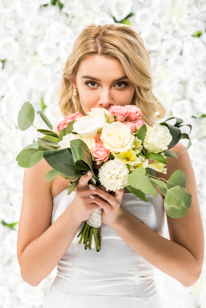 attractive young bride holding wedding bouquet near face on white floral background - Foto, Imagem