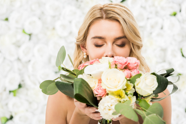 selective focus of young bride with closed eyes enjoying flavor of wedding bouquet on white floral background - Photo, Image